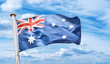 Australian flag blows in the wind against blue cloudy sky backgroundの写真素材