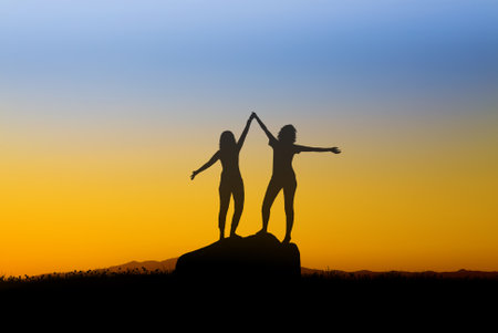 Two silhouetted girls raise their hands high, interlocking their fingers against the canvas of a vivid yellow-blue evening skyの写真素材