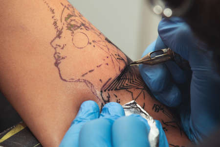 A tattoo artist paints a client's tattoo with ink. Tattoo artist holding a tattoo machine in Blue Sterile Gloves.の写真素材