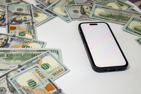 A smartphone with a blank screen is surrounded by scattered hundred-dollar bills on a clean white...