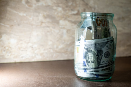Jar filled with hundred dollar bills placed on a wooden surface in a simple, well-lit settingの写真素材