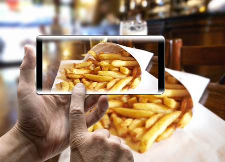 Two hands holding a mobile Smartphone and take a picture of Potatoes fries in a little white paper...