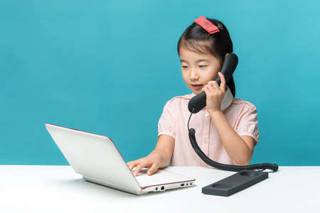 Cute asia little girl is sitting at table with her white laptop and a telephone, isolated over blue