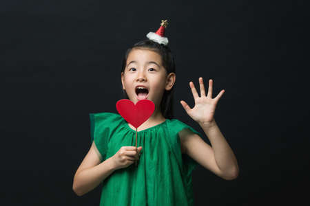 cute asian girl child dressed in a green dress holding a Christmas ornament and a heart stick on a black background. A surprised faceの写真素材