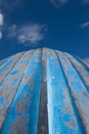 Hull of a blue weathered rowing boat, it's bow pointing up to a dark blue sky with white cloudsの写真素材