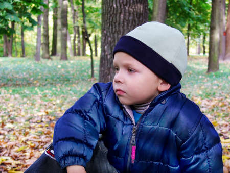 A little boy with an indifferent look sits on a bench in an autumn park. It is silence...の写真素材