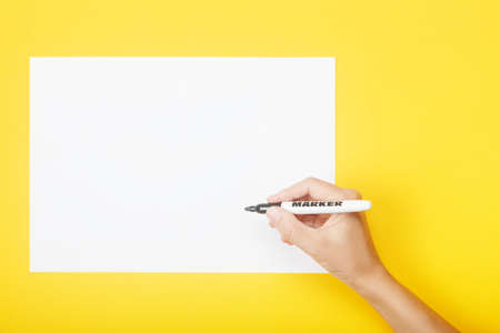 Hand with marker over blank paper on a yellow background. Mock up with copy space for your ideas. Flat lay style.の写真素材
