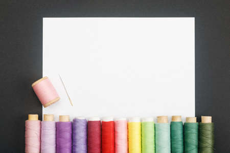 White sheet of paper on a black background with colored thread coils and a needle. The concept of sewing accessories. Layout with a copy of the space for your text. Flat layout, top view.の写真素材