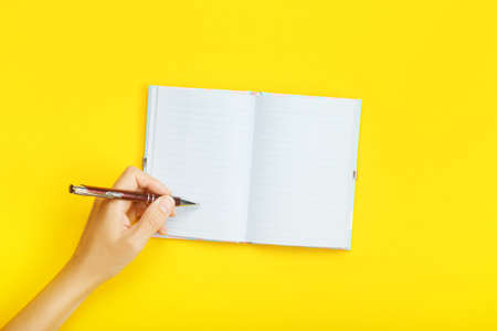 Hand with pen over blank notebook on a yellow background. Mock up with copy space for your ideas. Flat layout, top view.の写真素材