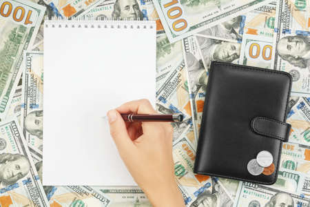Hand with ballpoint pen over blank notebook on background of dollar bills and coins on wallet....