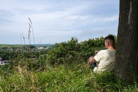 A young man sits under a tree overgrown with grass and looks into the distance, thinking about life, resting, relaxing. High quality photoの写真素材