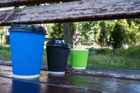 Three paper coffee cups with lids stand in a row on a wooden bench. Shot close-up, glasses of different sizesの写真素材