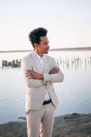A handsome dark-skinned African American man in a light suit with his bright red cabliolet car. The concept is a man-groom at sunset in a natural location outside the city. Emotion - fashion, seriouslyの写真素材