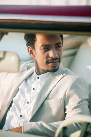 A handsome dark-skinned African American man in a light suit with his bright red cabliolet car. The...