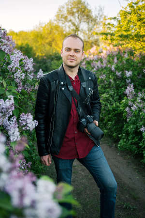Young male photographer with a camera in his hands against the background of lilac bushes in spring.