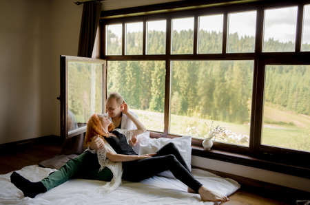 A beautiful couple in love hugs at the window, overlooking the mountains, in a wooden house, a romantic moment.の写真素材