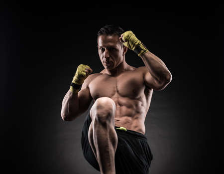 Muscular young man in boxing gloves and shorts shows the different movements and strikes in the studio on a dark backgroundの写真素材