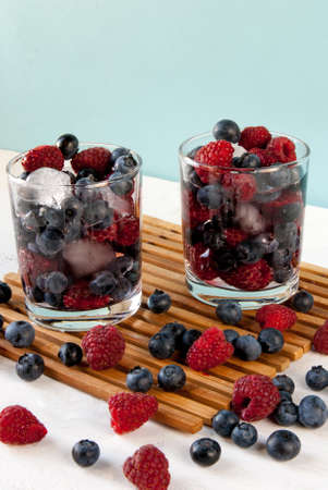 Cooled by ice juicy summer berries in glasses: raspberries, blueberries, bilberries. Rustic white table.の写真素材