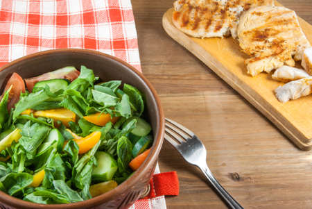 Chicken fillet, grilled, with a large portion of salad with fresh vegetables and herbs on a wooden table with a red cloth into a cell. top viewの写真素材
