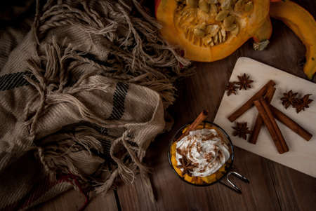Concept: fragrant autumn drink - all you need for pumpkin coffee latte with whipped cream and spices - aniseed and cinnamon. On the wooden background, next to a blanket, spices and pumpkinの写真素材