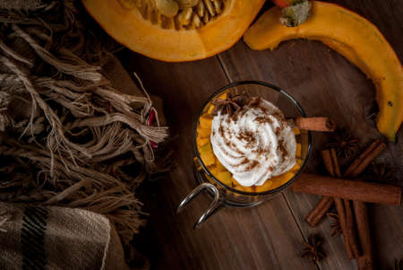 Concept: fragrant autumn drink - all you need for pumpkin coffee latte with whipped cream and spices - aniseed and cinnamon. On the wooden background, next to a blanket, spices and pumpkinの写真素材