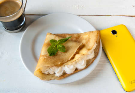 A light breakfast or lunch, with pancakes (crepes) and whipped cream. On the table is the coffee and smartphone. Sunny morning, white wooden tableの写真素材