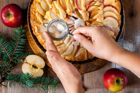 Woman decorates apple pie with powdered sugar. Wooden table, next to apples and branches of a Christmas tree and apples, top view, Hands in pictureの写真素材