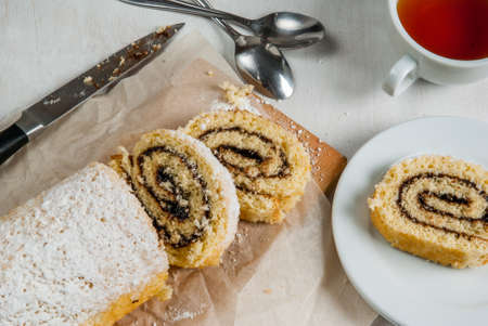 Biscuit roll with chocolate cream inside and powdered sugar on top. Roll is sliced, the next tea cup and plate with one slice of dessert. On a white wooden table, top viewの写真素材