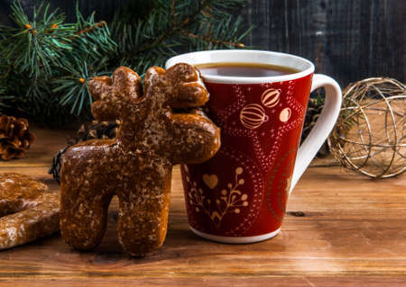A cup of tea and gingerbread in the form of a deer on a wooden table. Near the Christmas tree branch, pine cones and Christmas balls.の写真素材