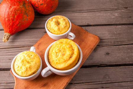 Pumpkin mug-cakes in rustic style, on an old wooden table, next to the cut and whole pumpkins and...