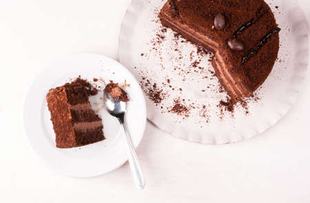 Chocolate cake with cream on a white wooden table. One piece of cake is on the plate, girl eats it, hand in the picture, top view, copy spaceの写真素材