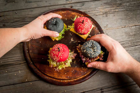 Small American colored burgers on a rustic wooden table, a snack at one bite. Man and woman take two burgers from board, hands in the picture, top viewの写真素材