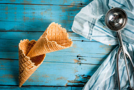 Traditional waffle cones for ice cream and a spoon for it on blue wooden table. Summer, open space, bright sun Wafers and chocolate chips in a frame on the table. One of cones filled with ice creamの写真素材
