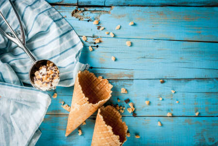 Traditional waffle cones for ice cream and a spoon for it on blue wooden table. Summer, open space, bright sun Wafers and chocolate chips in a frame on the table. One of cones filled with ice creamの写真素材