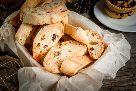 Traditional Christmas baking biscotti or cantucci in a basket on a wooden table next to the branches of a fir-tree, decorations and other kinds of Christmas sweets (tarts with nuts, ginger biscuits)の写真素材