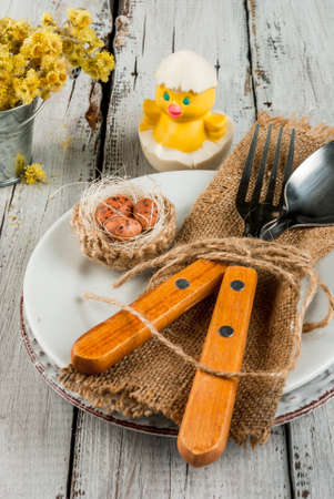 Spring Easter white wooden table setting with plate, decorative nest with egg, flowers, cutlery and...