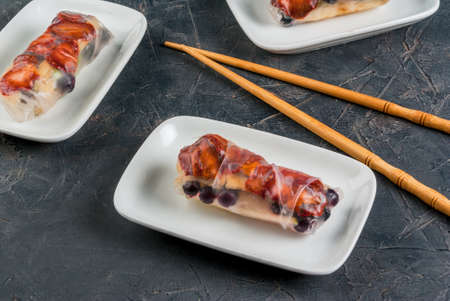 Sweet spring rolls with fruits and berries on the black stone table, next to chopsticks for sushi, close view, copy spaceの写真素材