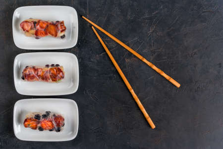 Sweet spring rolls with fruits and berries on the black stone table, next to chopsticks for sushi, top view, copy spaceの写真素材