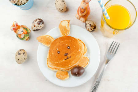 Cute and fun for children at Easter Breakfast: Pancake made in the shape of a bunny with chocolate egg in the legs. On a white table, with a glass of juice, surrounded by toys & decorations. Top viewの写真素材