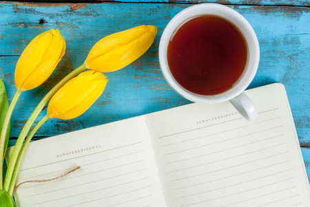 Tea mug with flowers yellow tulips and notebook for notes or wishing on blue rustic table top view, copy space, for Valentine's, Mothers day or Women's dayの写真素材