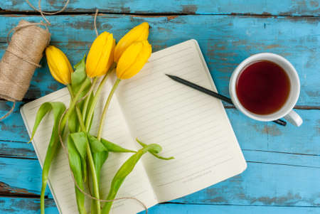 Tea mug with flowers yellow tulips and notebook for notes or wishing on blue rustic table top view, copy space, for Valentine's, Mothers day or Women's dayの写真素材