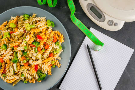 Concept of dieting, counting calories: a plate of healthy food (whole grain pasta with vegetables), scales for weighing food, tape measure, notepad and pencil for a food diary, top view, copy spaceの写真素材