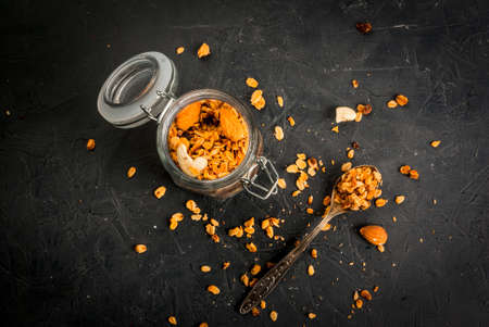 Golden granola cereal flakes with dried fruits, nuts and honey in a glass jar. On a dark stone...