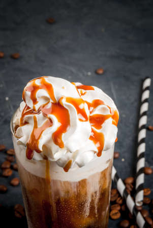 Cold coffee drink frappe (frappuccino), with whipped cream and caramel syrup, with straws and grains of coffee on a dark gray stone table, copy spaceの写真素材