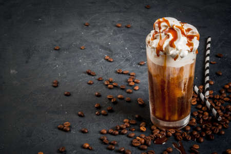 Cold coffee drink frappe (frappuccino), with whipped cream and caramel syrup, with straws and grains of coffee on a dark gray stone table, copy spaceの写真素材
