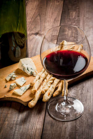 Red wine and snacks for it: Italian grissini bread sticks and blue cheese on a cutting wooden board on a wooden table, copy spaceの写真素材