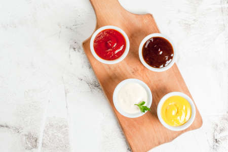 Classic set of sauces in white saucers: American yellow mustard, ketchup, barbecue sauce, mayonnaise. On cutting board white stone concrete table top view, copy spaceの写真素材