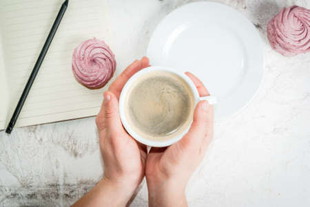 Home spring vacation, relax, work. White stone concrete desk with notepads, coffee and pink berry pastries (marshmallows). The girl holds a cup in his hands top view copy spaceの写真素材