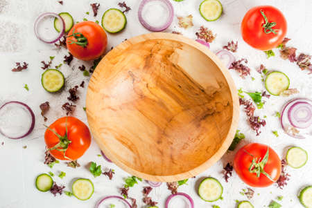 Spring salad concept: fresh lettuce, onion rings, parsley, greens, cucumbers and tomatoes on a white table, with a wooden bowl of salad. Flat top view, background with ingredients, copy spaceの写真素材
