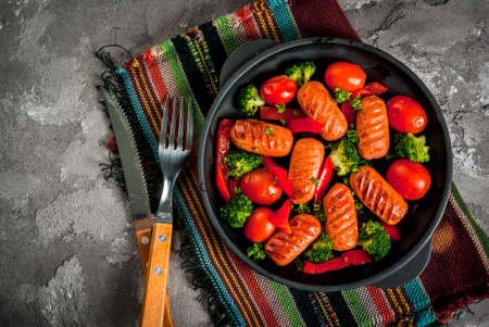 Grilled sausages and vegetables (tomatoes, broccoli, pepper) in a frying pan on a concrete gray table, top view, copy spaceの写真素材